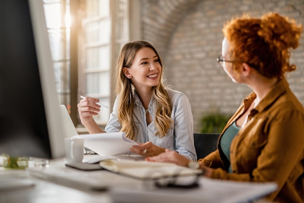 Smiling creative businesswoman going through paperwork and talking to her female colleague in the office