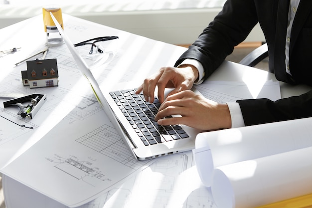 Hands of CEO of construction company in black suit typing message via e-mail to partners on laptop with drawings, seal stamp, divider on table.